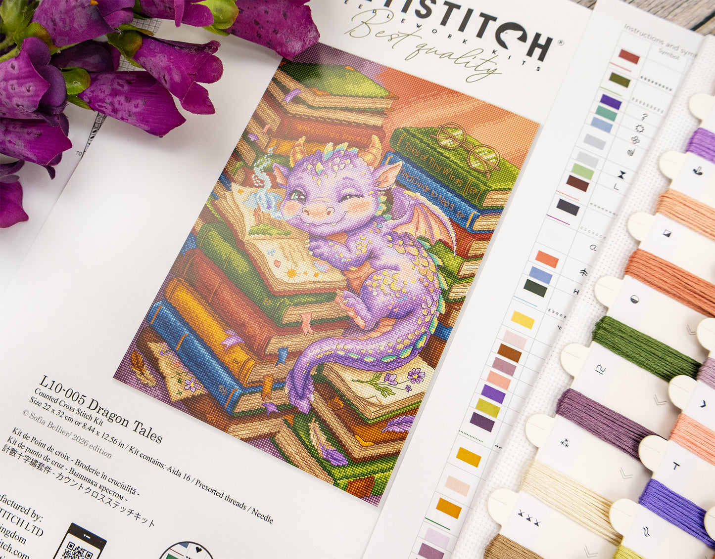 A counted cross-stitch pattern booklet titled 'Dragon Tales' is shown open, featuring a colorful illustration of a purple dragon resting on stacked books. The booklet is styled on a white surface with purple flowers on the left and a thread color sampler card on the right.