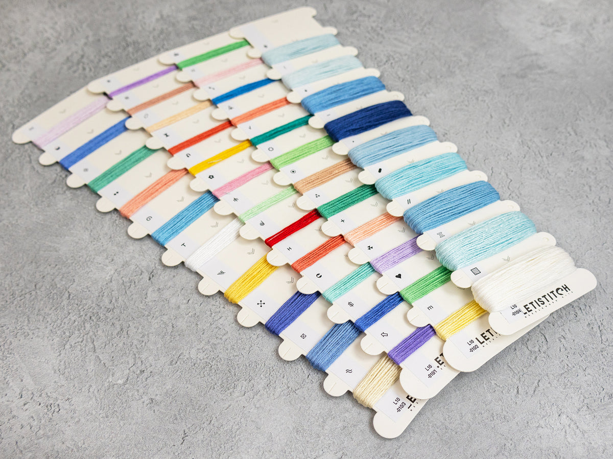 A fan-shaped arrangement of many embroidery floss skeins in assorted colors on a light gray textured background. The labels show the brand Letistitch.