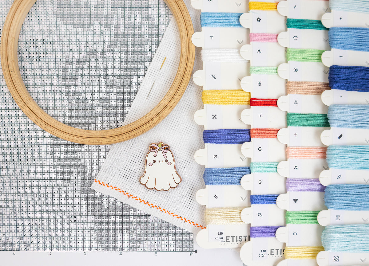 A cross-stitch kit setup featuring a wooden hoop with white thread, a white fabric canvas with a gray pattern grid, a small ghost-shaped wooden ornament, and multiple spools of colorful embroidery thread on the right side.