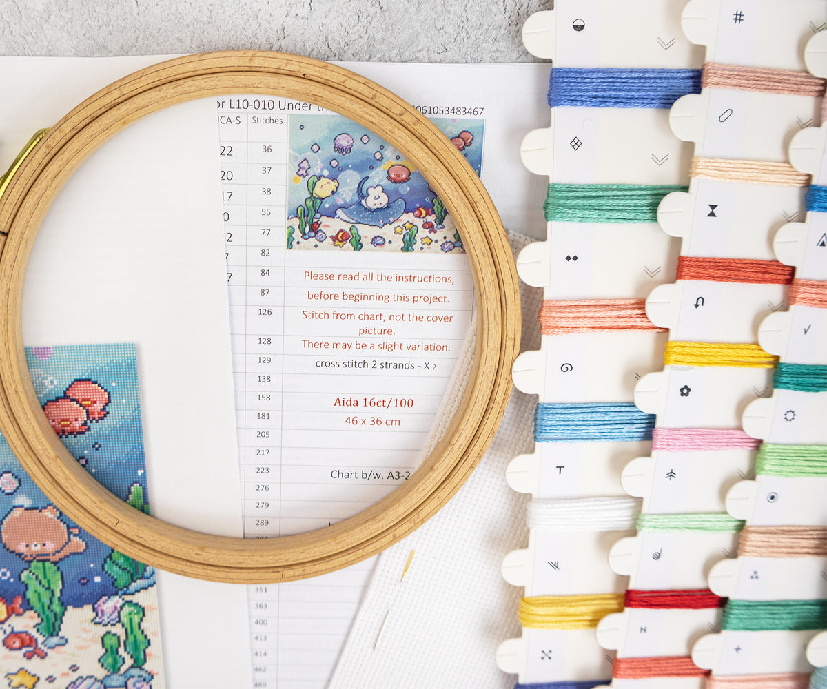 A wooden embroidery hoop holding a printed chart and colorful underwater-themed embroidery design, shown next to a rack of multicolored embroidery floss threads on a light surface.