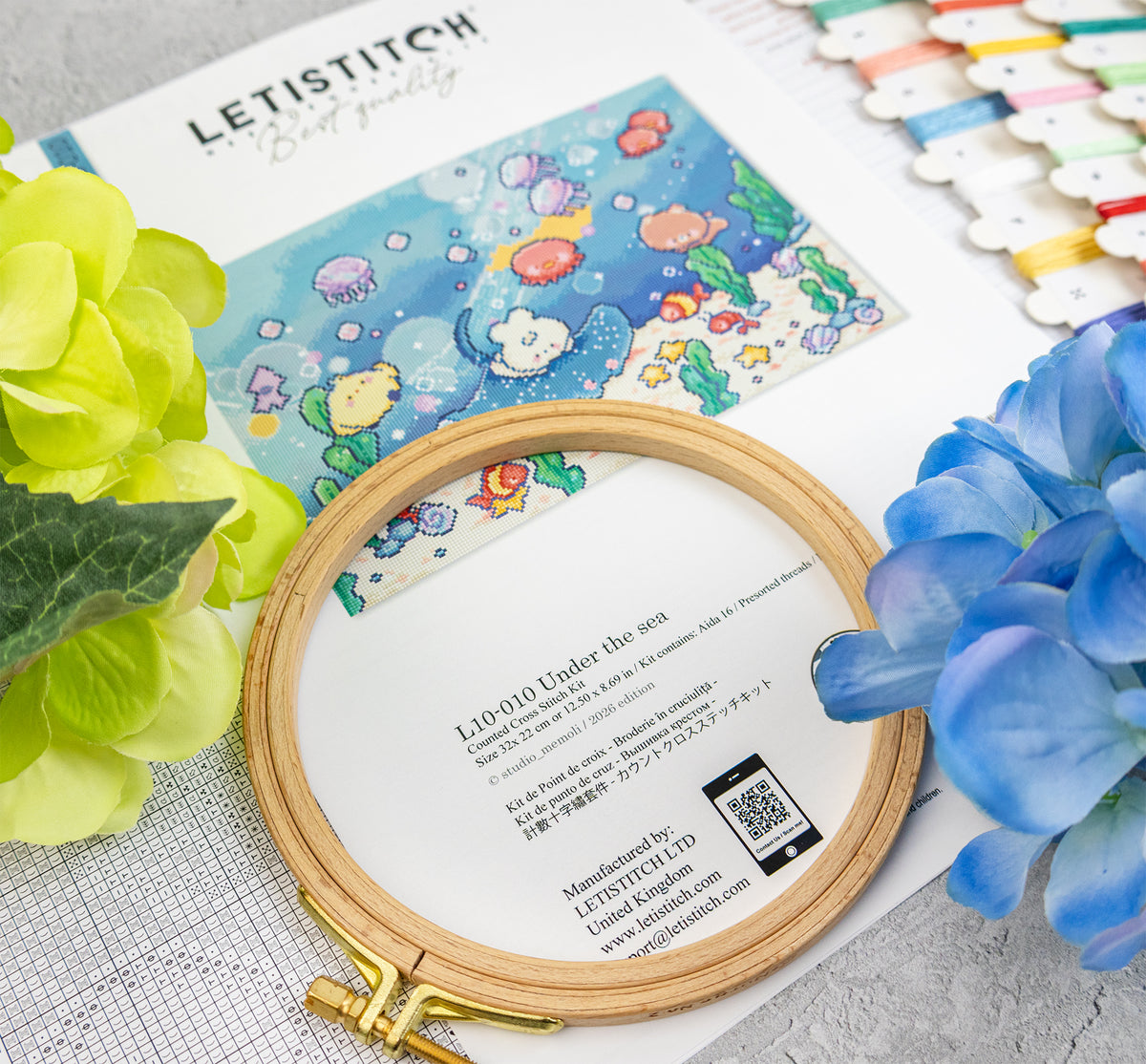 A wooden embroidery hoop holding white thread, placed over a printed Letistitch pattern sheet with an underwater design. The scene includes decorative flowers, thread spools, and a textured surface, with the Letistitch brand name visible on the sheet and packaging.