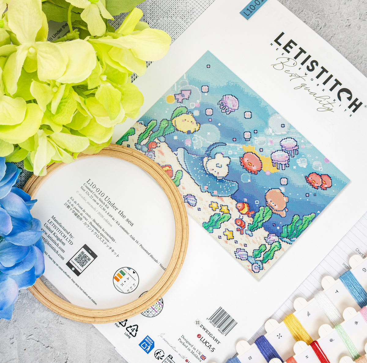 A flat-lay of a cross-stitch kit featuring the design 'Under the sea' by Letistitch. The white paper sheet displays a colorful underwater scene with a turtle, fish, and sea plants, alongside a wooden embroidery hoop, thread spools, and decorative green and blue flowers on a light gray textured surface.