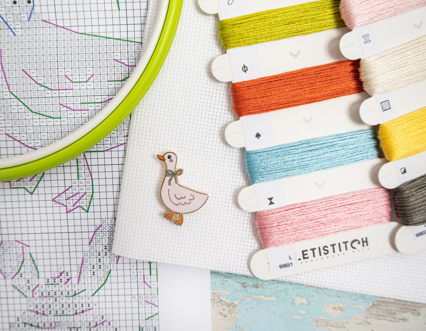 Embroidery hoop with colorful threads and a small duck charm on a grid background
