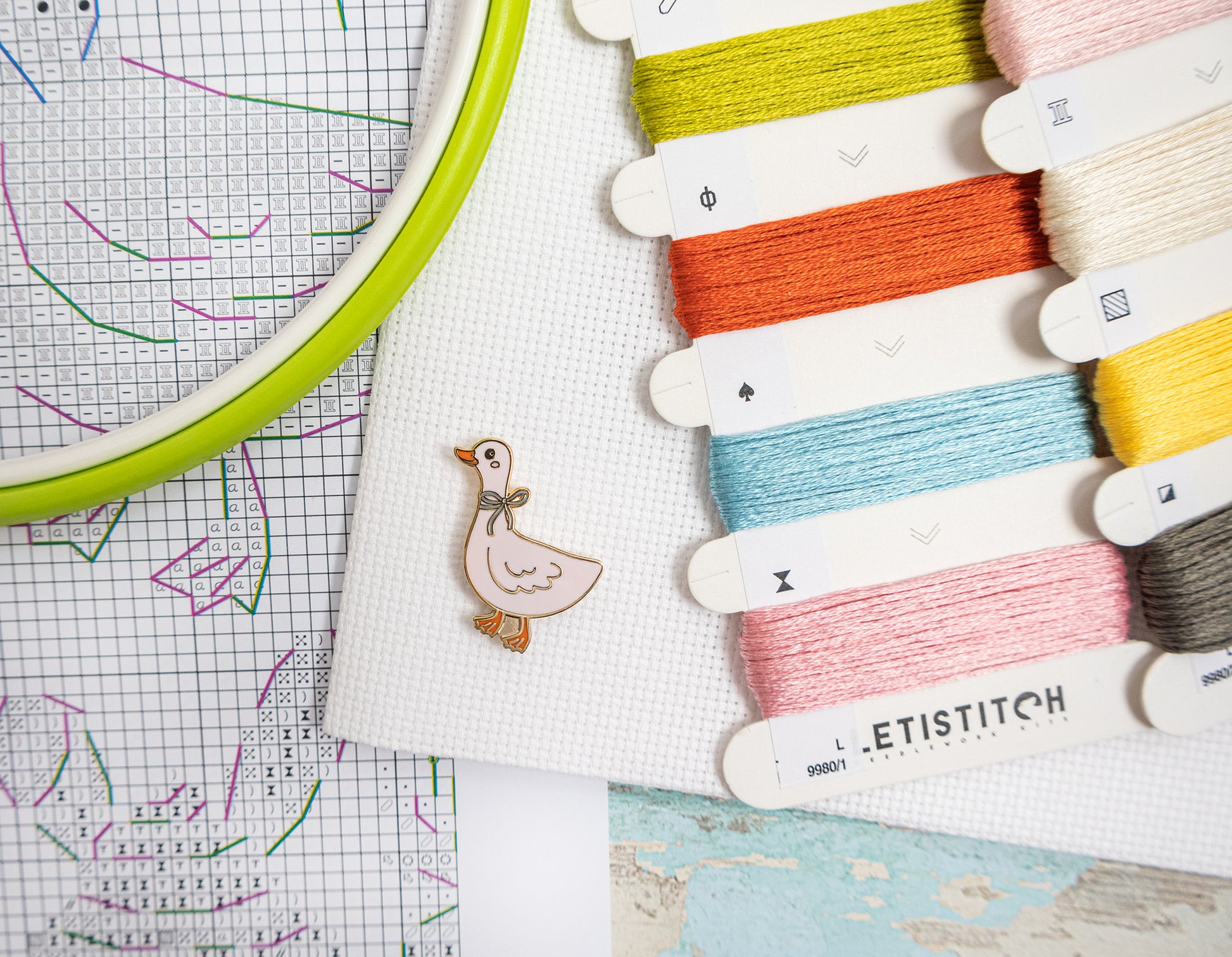 Embroidery hoop with colorful threads and a small duck charm on a grid background
