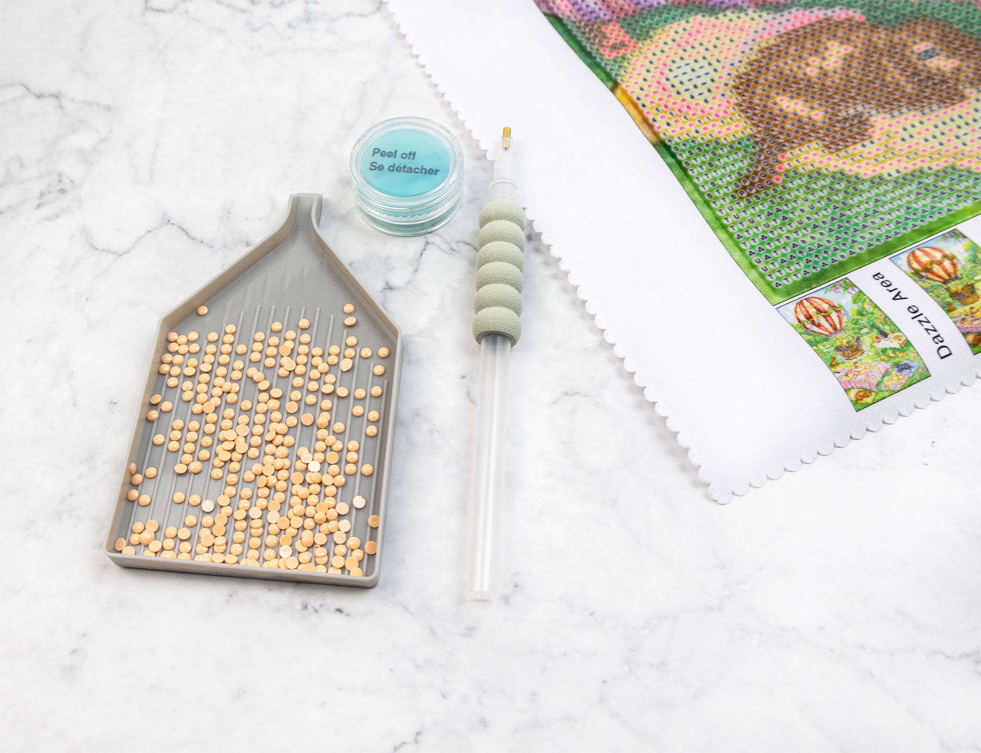 Crafting materials including crystals a tool, and a pattern sheet on a white surface for Diamond Painting 